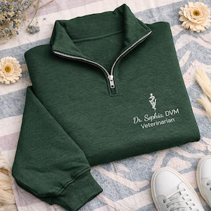 May include: A dark forest green quarter-zip sweatshirt featuring a silver zipper and ribbed cuffs. Embroidered in white on the chest is a veterinary caduceus symbol above the text Dr. Sophia, DVM Veterinarian in a clean, professional font.