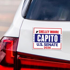 May include: A rectangular bumper sticker with a red and white border. The sticker has the words "Shelley Moore Capito" in red and blue, with "U.S. Senate 2026" below. The sticker is affixed to the rear of a white car.