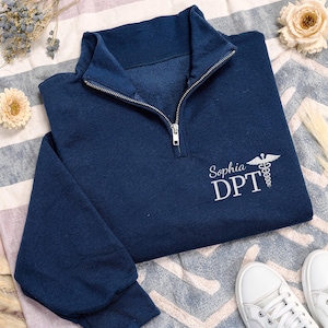 May include: A navy blue quarter-zip sweatshirt featuring a silver zipper and white embroidered text on the chest that reads Sophia DPT, accompanied by a medical caduceus symbol. The garment has long sleeves and a high collar design.