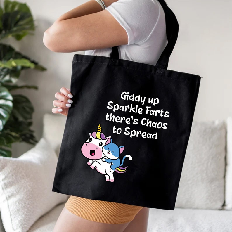 May include: Black tote bag with a white unicorn and a blue cat riding on its back. The bag has the text "Giddy up Sparkle Farts there's Chaos to Spread" printed on it.