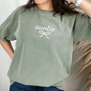 May include: Sage green t-shirt with the word "auntie" in a cursive font and a bow graphic. The shirt has short sleeves and a relaxed fit. The text and graphic are in white.