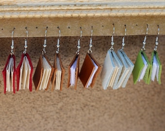 Puede incluir: Un conjunto de pendientes hechos a mano con forma de libro en varios colores, incluyendo rojo, marrón, azul claro y verde. Cada pendiente presenta un diseño de libro pequeño y abierto con páginas detalladas y ganchos plateados. Los pendientes se exhiben en un estante de madera.