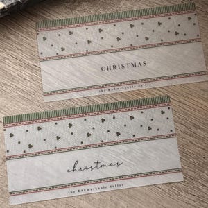 May include: Two rectangular paper slips featuring a light gray background with a subtle wood grain texture. Each slip displays a green and red patterned border, the word CHRISTMAS or christmas in black text, and the phrase the RAEmarkable dollar.
