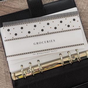 Puede incluir: Un archivador de cuero sintético negro con anillos dorados, abierto para revelar un bolsillo transparente con la etiqueta "GROCERIES". El bolsillo tiene un estampado floral y el texto "the RAEmarkable dollar". Se ve una tarjeta con la palabra "intentional".