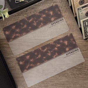 May include: Two rectangular cards with a tortoiseshell pattern and the word "SPENDING" printed on them. The cards have a light gray section at the bottom. The cards are on a wooden surface, with cash and a black tray with the word "intentional" visible.