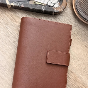 May include: A brown, textured, rectangular personal planner with a cross-hatched finish and a matching strap closure. The cover features a vertical spine and a small pen loop on the right side, presenting a clean, structured design for daily organization.