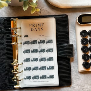 May include: A white sheet of paper with the text "PRIME DAYS" and "GOAL" printed on it. There are 20 black and white stickers of Amazon delivery trucks with the word "prime" printed on them. The paper is in a black binder with gold rings.
