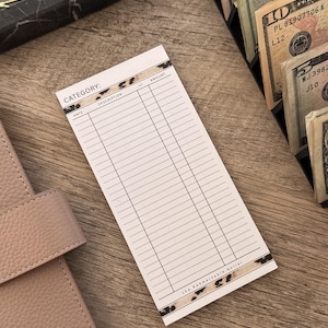 May include: A white ledger sheet with a leopard print border, featuring columns for date, description, and amount. The text "CATEGORY:" is visible at the top. The ledger is on a wooden surface, near a pink leather notebook and a cash organizer.