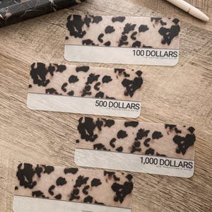 May include: Four gift cards with a cow print design. The cards are labeled with the amounts: 100, 500, and 1,000 dollars. The cards have a white section at the bottom with the text "the remarkable dollar". A pen and a marble tray are also in the image.