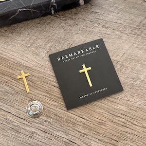 May include: A gold-colored cross pin and a silver magnetic backing are displayed on a wooden surface. The cross is also shown on a black card with the text "RAEMARKABLE EVERY DETAIL, ON PURPOSE MAGNETIC ACCESSORY."