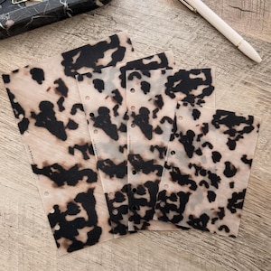 Puede incluir: Insertos de planificador translúcidos con un diseño de carey en tonos marrón y negro. Los insertos están perforados para encuadernar y descansan sobre una superficie de madera. Un bolígrafo y una bandeja decorativa son visibles.