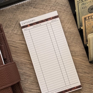 May include: A white notepad with a brown tortoiseshell border, featuring the text "CATEGORY:" and columns for date, description, and amount. The notepad is on a wooden surface, with a brown leather wallet and cash visible.