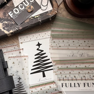 May include: A collection of translucent plastic cash envelopes for a binder, featuring holiday-themed designs including a black Christmas tree graphic, green and red patterned borders, and printed text such as "Christmas," "FULLY FUNDED," and "100," "500," "1,000."