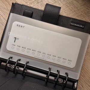 May include: A black six-ring binder featuring a translucent plastic divider with the text RENT, 1st, and the months JAN through DEC with corresponding circular checkboxes. The brand name RAEMARKABLE is printed on the divider and on a small black tab.