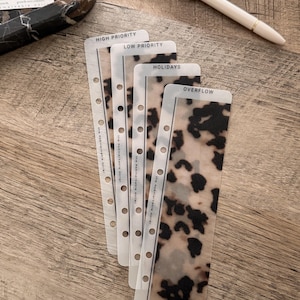 May include: Planner dividers with a translucent leopard print pattern. Each divider is labelled with text: "HIGH PRIORITY", "LOW PRIORITY", "HOLIDAYS", and "OVERFLOW". A gold-toned pen rests on a wooden surface.