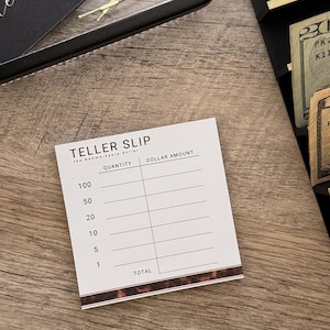 Pode incluir: Um bloco de notas branco "TELLER SLIP" com uma borda marrom, com seções de quantidade e valor em dólares. O bloco de notas está em uma superfície de madeira, com uma bandeja preta e dinheiro visível no fundo.