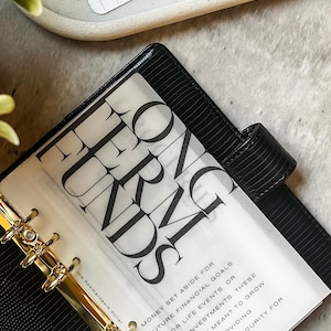 May include: A black leather planner with gold rings and a clear plastic sheet with the words "LONG TERM FUNDS" printed in black. The sheet also includes text about setting aside money for future financial goals, major life events, and large investments.