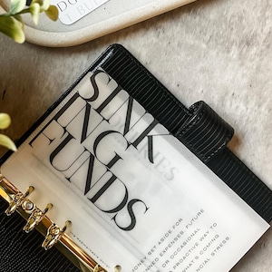 May include: A black leather planner with gold rings and a white sheet of paper with the words "Sink Funds" and the text "Money set aside for planned expenses, future needs or occasional costs. A proactive way to save for what's coming without financial stress." printed on it.