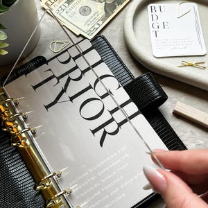 May include: A black planner with gold accents, open to a page with the word "PRIOR" in large, bold letters. The planner is next to a tray with a "BUDGET" card, cash, and a small plant.