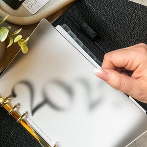 Op de afbeelding: Een zwarte lederen planner met gouden ringen en een witte plastic insert met de maanden van het jaar gelabeld. De planner is open naar de sectie 2022.