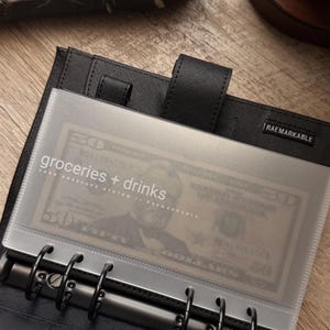 May include: A black six-ring binder featuring a translucent plastic cash envelope insert labelled groceries + drinks. The binder has a textured exterior, a metal ring mechanism, and a small black tag with the brand name RAEMARKABLE attached.