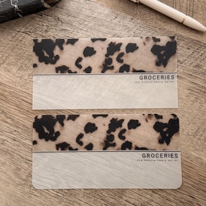 May include: Two rectangular cards with a tan and black spotted pattern on top and a white section below. The text "GROCERIES the remarkable dollar" is printed on the white section. A pen and a small decorative tray are in the background.