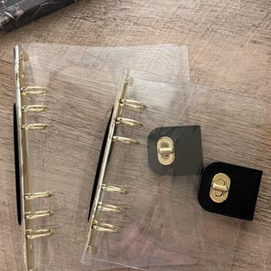May include: Clear planner with gold-colored metal rings and black leather clasp. The planner is open, revealing the rings and clear plastic pages. The clasp is closed with a gold-colored metal lock.