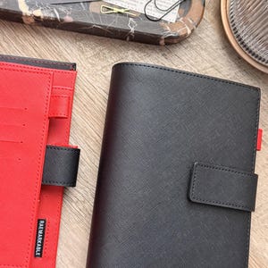 May include: A black textured planner cover with a magnetic strap closure, positioned next to an open red planner cover featuring a black pen loop and a small black tag with the white text RAE MARKABLE printed vertically on the lower edge.