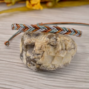 May include: Braided leather bracelet in shades of gray, teal, and brown. The bracelet is displayed on a textured rock. The adjustable cord is teal and brown. The bracelet is a stylish accessory.