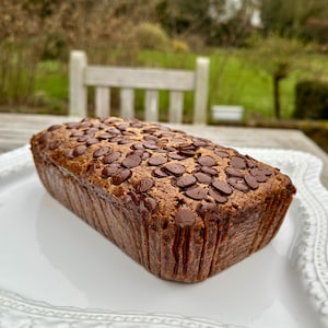 May include: A loaf of chocolate chip banana bread on a white plate. The bread is a light brown colour and has a slightly crispy crust. The chocolate chips are evenly distributed throughout the bread.