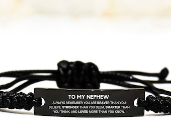May include: Black braided bracelet with a rectangular metal plate engraved with the message "TO MY NEPHEW". The text reads "ALWAYS REMEMBER YOU ARE BRAVER THAN YOU BELIEVE, STRONGER THAN YOU SEEM, SMARTER THAN YOU THINK, AND LOVED MORE THAN YOU KNOW."