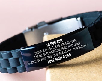 Op de afbeelding: Zwart siliconen armband met een zilveren metalen plaat gegraveerd met de boodschap "Aan onze zoon, moed is niet de afwezigheid van angst, maar de vastberadenheid om iets te doen ondanks de angst. Heb de moed om je dromen na te jagen. Liefde van mama en papa."
