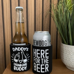 May include: Two black beverage insulators with white text. One insulator says "Daddy's Drinking Buddy" with a baby bottle and beer bottle graphic. The other insulator says "Here for the Beer".