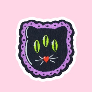 May include: Black cat embroidered patch with green eyes, a red heart nose, and white whiskers. The patch has a purple lace trim and is surrounded by a white border.