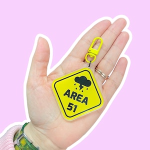 May include: A yellow Area 51 keychain with a black cloud and lightning bolt graphic. The keychain has a yellow clasp and a clear plastic backing. The text "AREA 51" is printed in black on the yellow background.