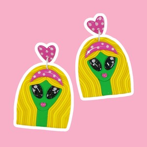 Op de afbeelding: Een paar oorbellen met een groen alien gezicht met roze stippen hoofdbanden en roze hart accenten. De alien gezichten hebben grote ogen en een roze mond.