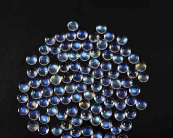 Op de afbeelding: Een verzameling van talrijke kleine, ronde, doorschijnende cabochons met een glad, gewelfd oppervlak. Elk stuk vertoont een uitgesproken iriserende blauwe gloed, kenmerkend voor maansteen, verspreid over het kader in een losse, cirkelvormige opstelling.