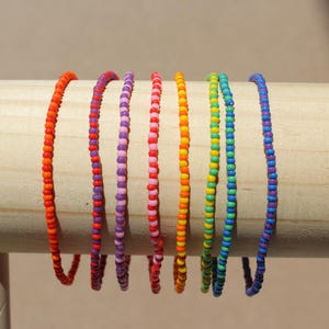 May include: Seven colourful beaded bracelets displayed on a wooden dowel. The bracelets feature a spectrum of colours: red, orange, purple, pink, yellow, green, and blue. Each bracelet is approximately 17.78 cm long, showcasing a delicate, vibrant design.
