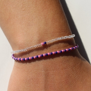Peut inclure: Deux bracelets en perles sur un poignet. Le bracelet du haut est fait de perles transparentes. Le bracelet du bas est fait de perles roses et violettes.