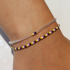 Peut inclure: Deux bracelets en perles sur un poignet. Un bracelet est transparent et l'autre est violet, jaune, blanc et noir.