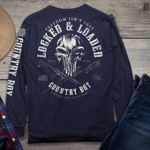 Op de afbeelding: Een marineblauw shirt met lange mouwen met een witte schedelprint en de tekst "Freedom Isn't Free Locked & Loaded Country Boy American Original 1776". De mouwen hebben de tekst "Country Boy" verticaal gedrukt.