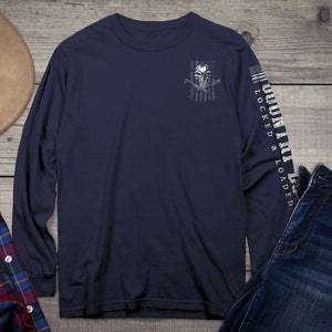 Op de afbeelding: Een marineblauw longsleeve t-shirt met een witte schedel en gekruiste botten afbeelding op de voorkant. De afbeelding is geplaatst tegen een vervaagde Amerikaanse vlag ontwerp. De rechtermouw heeft de tekst "COUNTRY LOCKED & LOADED" in wit.