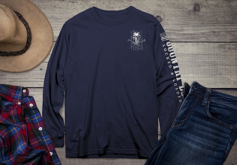 Op de afbeelding: Een marineblauw longsleeve t-shirt met een witte schedel en gekruiste botten afbeelding op de voorkant. De afbeelding is geplaatst tegen een vervaagde Amerikaanse vlag ontwerp. De rechtermouw heeft de tekst "COUNTRY LOCKED & LOADED" in wit.