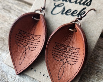 May include: A pair of brown leather earrings with a geometric design. The earrings are on a brown paper card with the text "Cactus Creek" written in black.