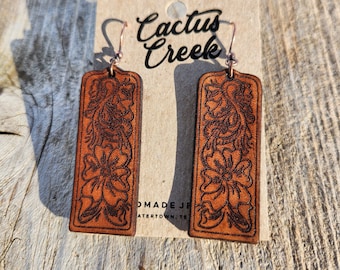 May include: A pair of rectangular brown leather earrings featuring a dark embossed floral pattern and copper-toned metal hooks. The earrings are attached to a tan cardboard display card printed with the text Cactus Creek.