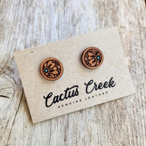 May include: Two round, genuine leather earrings with a floral design and a turquoise center stone. The earrings are on a tan card with the words "Cactus Creek Genuine Leather" in black script.
