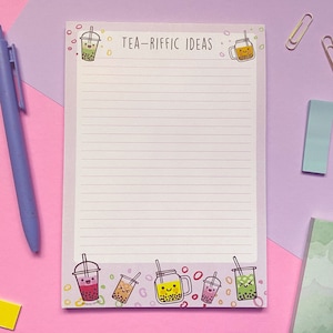 May include: A pink and white notepad with a lined interior and a whimsical design featuring cartoon teacups with smiling faces. The notepad has the text "TEA-RIFFIC IDEAS" at the top.
