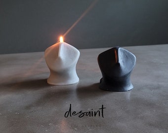 May include: Two sculptural candles, one lit, in a minimalist design. The lit candle is white, while the other is black. The candles have a unique shape and are placed on a gray surface. The word "desaint" is written in cursive.