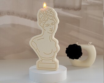 May include: A cream-colored candle shaped like a classical bust, with a golden outline. The candle is on a white pedestal, with a small, round candle and a black accent piece in the background. The candle has a faux flame.