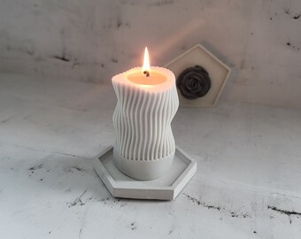 May include: A lit, gray, wavy-patterned candle sits on a hexagonal gray tray. The candle has a unique, textured design and is burning with a bright flame. A small, decorative rose is in the background.